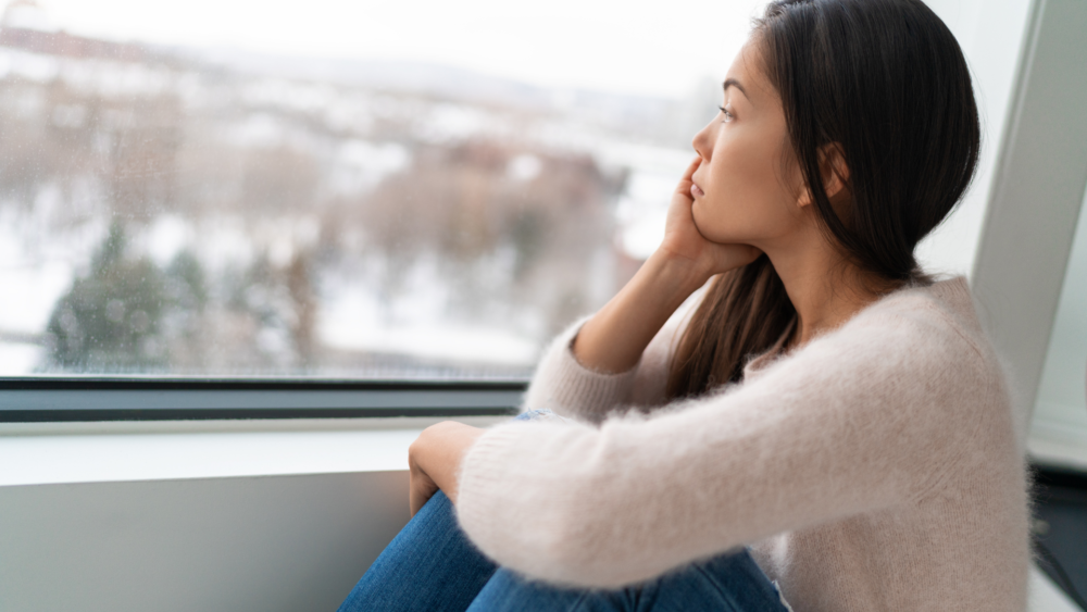 Mujer triste mirando por una ventana con expresión de melancolía, representando la duda emocional en una relación. Reflexión sobre el amor y la conexión en pareja, disponible en Micentrolasrozas.es. | Apego ansioso en parejas