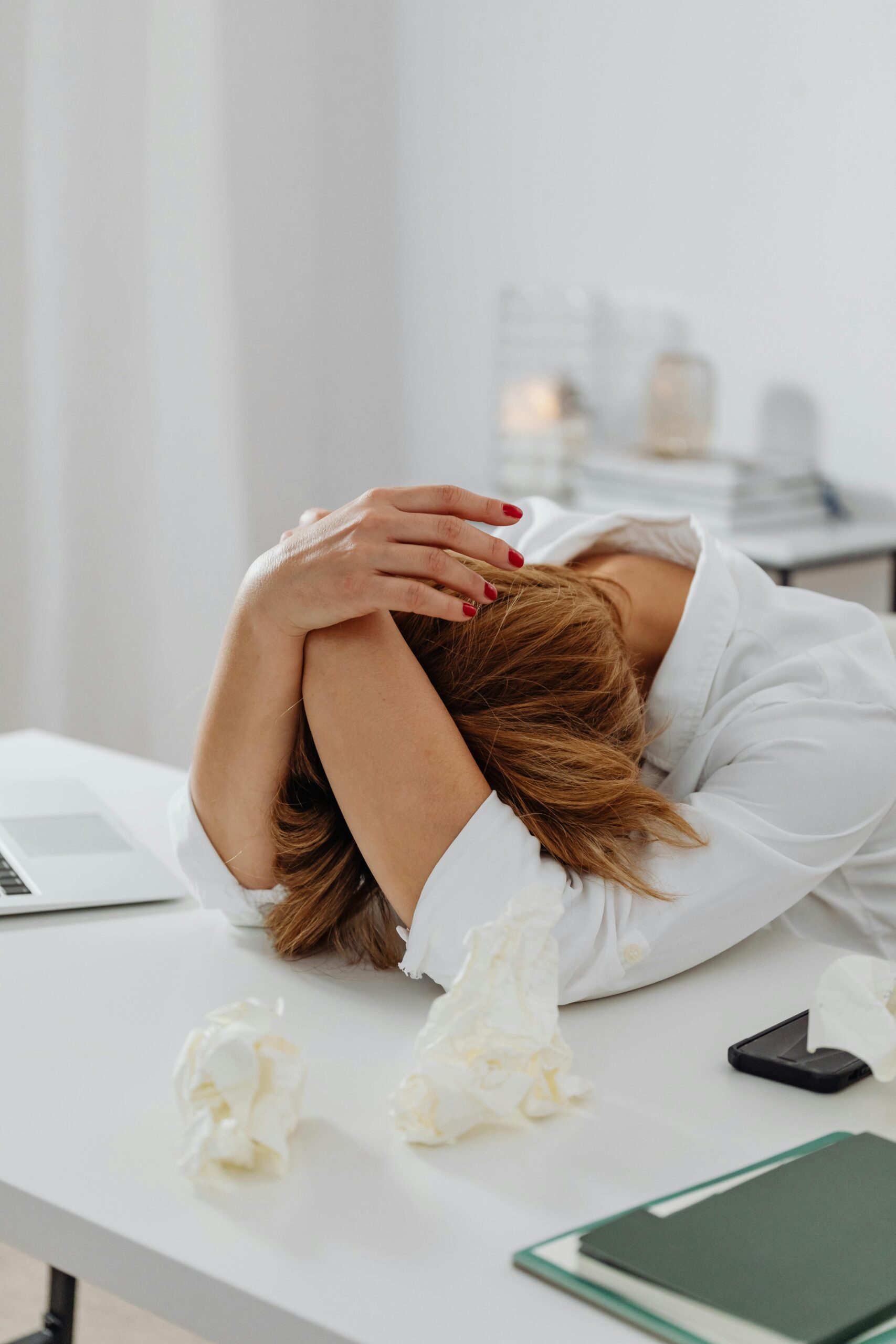 Mujer con la cabeza apoyada en el escritorio, visiblemente cansada, rodeada de papeles arrugados, mostrando signos de fatiga profunda asociada al estrés celular